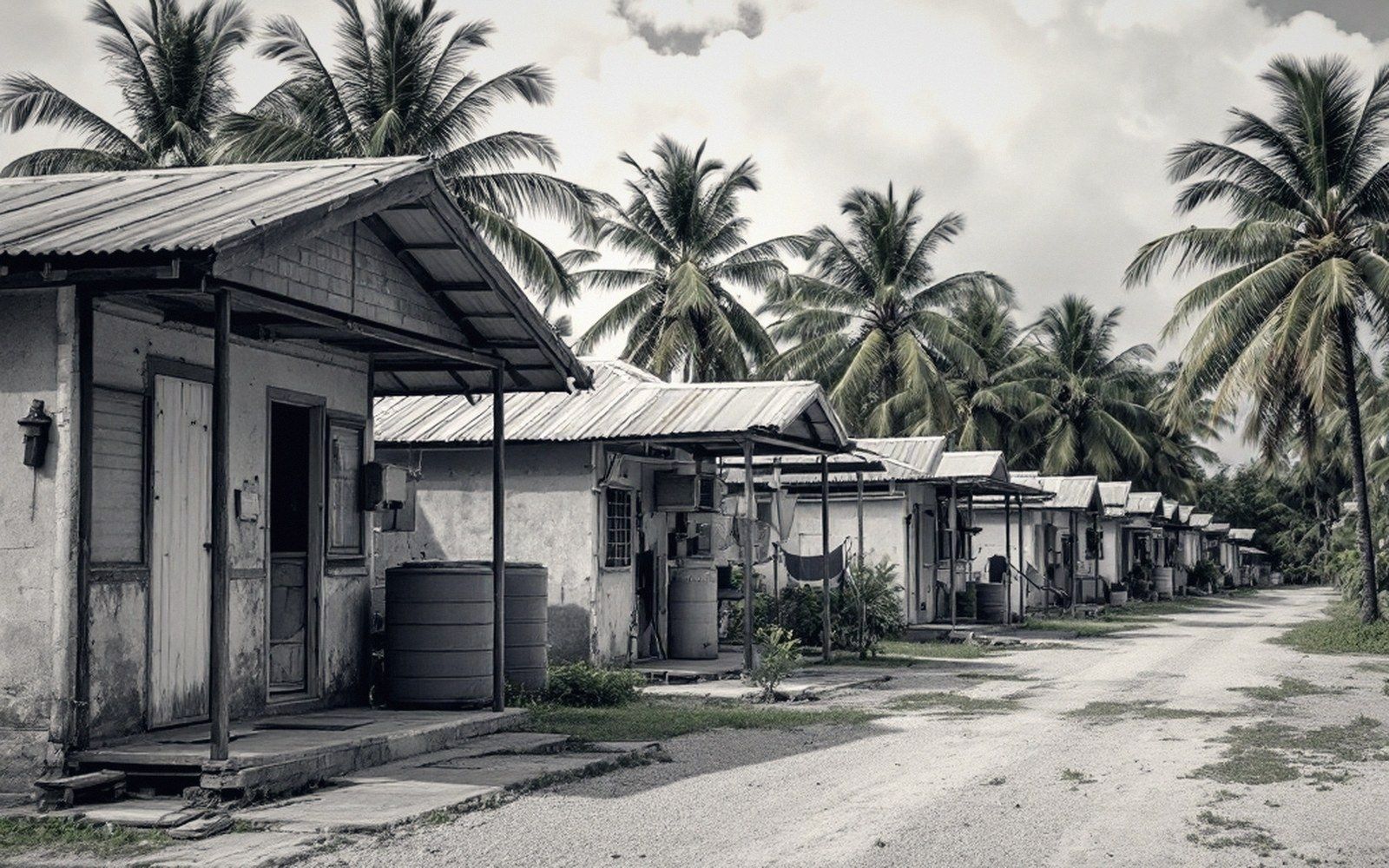 A row of tin-roofed cottages with water barrels lines a dirt path under palm trees